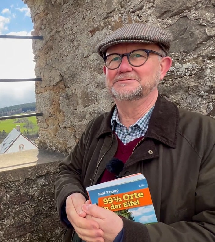 Ralf Kramp vor einer Mauer und hält sein Buch in der Hand