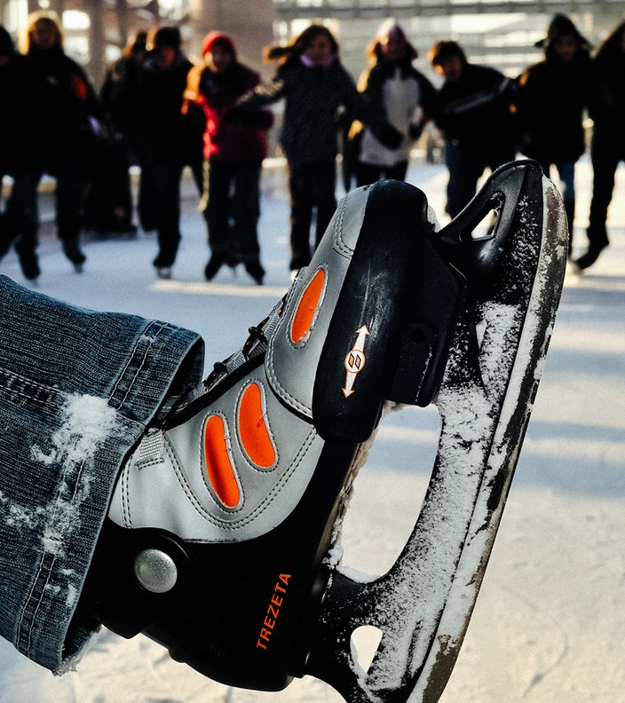 Ein Fuß mit einem Schlittschuh, im Hintergrund weitere Menschen mit Schlittschuhen und die Zeche Zollverein