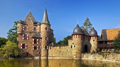 An einem Teich gelegen eine Burg aus braunem Stein mit grauem Dach. Ein Torhaus mit zwei Türmen.