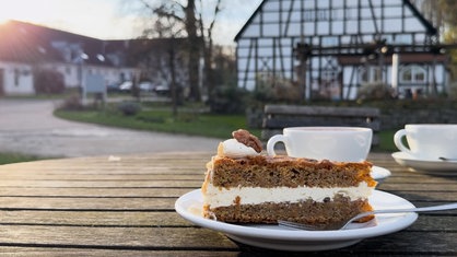 Ein Stück Kuchen auf einem Teller in Nahaufnahme. Im Hintergrund der Bauernhof Schultenhof.