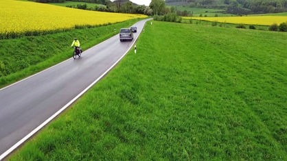 Eine schmale Straße zwischen gelbem Rapsfeld und grüner Wiese, darauf ein Fahrradfahrer und zwei Autos.