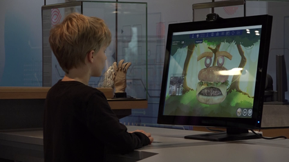 Auf dem Bild sieht man einen Computerbildschirm. Dieser zeigt ein Spiel an. Vor dem Bildschirm steht ein kleiner Junge, der das Spiel spielt.