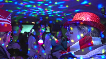 Der Innenraum des Taxis wird durch Party-Lichter erhellt. Im Vordergrund eine Frau mit rot-weißem Schal und einem roten Hut.