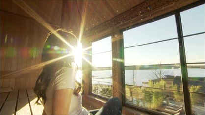 Frau sitzt angezogen in einer Holzsauna und blick durch Panoramafenster auf einen See