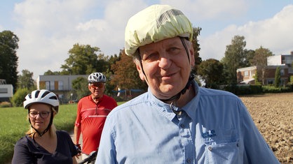 Älterer Mann in hellblaumen Hemd mit Helm auf dem Kopf steht in der Natur und spricht in Kamera, im Hintergrund weitere Fahrradfahrer