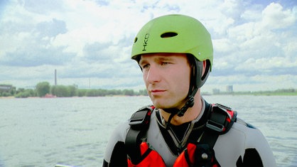 Ein Mann trägt einen Schwimmanzug und grünen Helm. Im Hintergrund ist ein Fluss zu sehen.