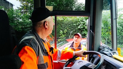 Zwei Männer in orangener Arbeitskleidung unterhalten sich. Einer von ihnen sitzt im Auto, der andere steht am Fahrerfenster