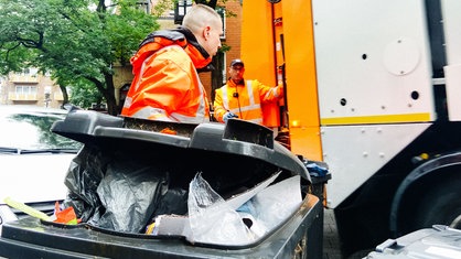 Ein Mann in neon-orangener Arbeitskleidung zieht eine übervolle Mülltonne Richtung Müllauto