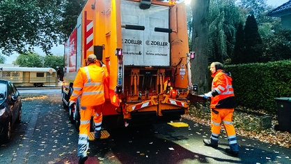 Zwei Männer in neon-orangener Arbeitskleidung steigen auf zwei Trittbretter eines Müllautos