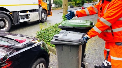 Ein Mann in neon-orangener Arbeitskleidung und Schutzhandschuhen zeigt auf ein schwarzes Auto, das zwei Mülltonnen zugeparkt hat