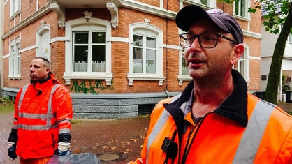 Ein Mann mit schwarzer Brille in orangener Arbeitskleidung und schwarzer Kappe auf dem Kopf