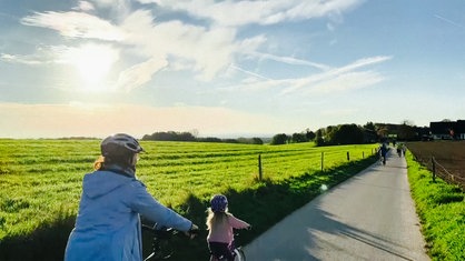 Ein grünes Feld. In der Mitte ein asphaltierter Weg auf dem sich einige Radfahrer in Richtung Horizont bewegen