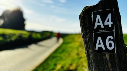 Ein brauner Pfahl mit weißen Wanderwegbeschriftungen. Im Hintergrund ein Weg umgeben von grünen Feldern