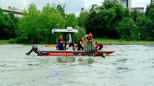 Ein Boot, auf dem mehrere Personen stehen. Eine Person wird mit einem Netz gerade aus dem Wasser gehoben.