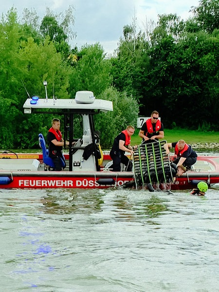 Ein Boot, auf dem mehrere Personen stehen. Eine Person wird mit einem Netz gerade aus dem Wasser gehoben.