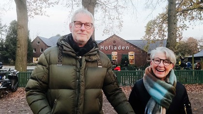 v. l. ein Mann und eine Frau. Sie stehen vor einem Hofladen. Beide lächeln.