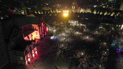 Der Platz vor dem beleuchteten Dom in Münster von oben, auf dem Platz stehen tausende Menschen und eine kleine Bühne