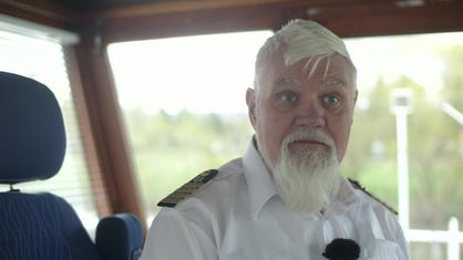 Mann mit weißen Haaren und weißem Bart trägt eine Kapitänsuniform und schaut in Kamera 