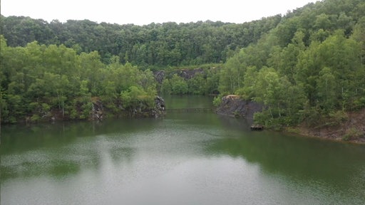 Stillgelegter Steinbruch im Naturschutzgebiet Schlupkothen. Der Steinbruch ist mit Wasser vollgelaufen, an den Rändern ragen Felswände empor, überall wachsen Sträucher und Bäume.