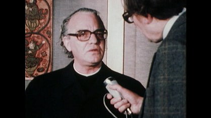 Mann mit grauem Haar und schwarzer Brille spricht in ein Mikrofon