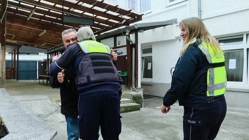 Ein Polizeibeamter in grün-blauer Uniform der einen Mann mit Bart umarmt. Neben ihm eine weitere Polizeibeamte