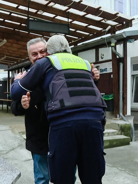 Ein Polizeibeamter in grün-blauer Uniform der einen Mann mit Bart umarmt. Neben ihm eine weitere Polizeibeamte