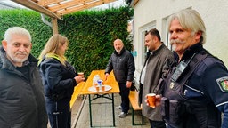 Zwei Männer mit grauen Haaren und Bart. Einer davon trägt eine Polizeiuniform. Im Hintergrund steht eine weitere Beamtin und zwei Männer