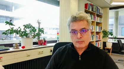 Ein Mann mit kurzen grauen Haaren und Brille sitzt in einem Büro mit einem großen Bücherregal im Hintergrund