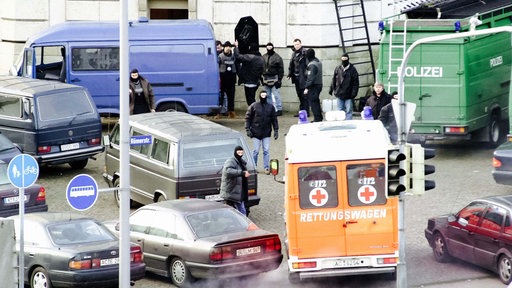 Chaos auf dem Vorplatz eines Gebäudes. Autos stehen durcheinander, Menschen laufen zwischen ihnen hindurch. Ein Krankenwagen. Ein Polizeifahrzeug.