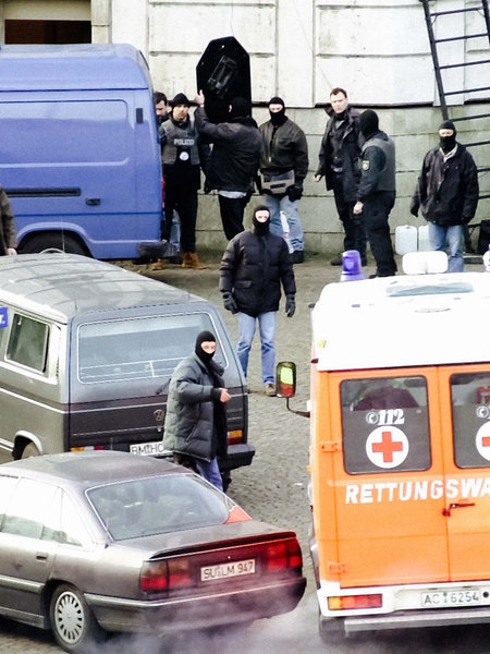 Chaos auf dem Vorplatz eines Gebäudes. Autos stehen durcheinander, Menschen laufen zwischen ihnen hindurch. Ein Krankenwagen. Ein Polizeifahrzeug.