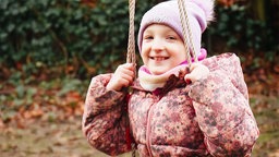 Melina, ein junges Mädchen in rosa Winterjacke und rosa Bommelmütze, sitzt auf einer Schaukel und lacht in die Kamera