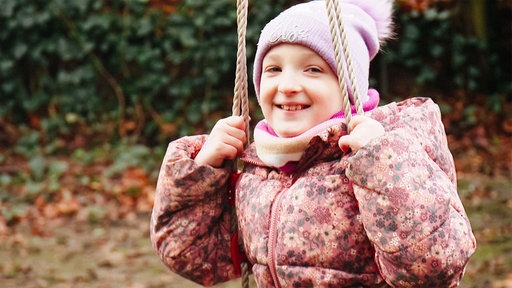 Melina, ein junges Mädchen in rosa Winterjacke und rosa Bommelmütze, sitzt auf einer Schaukel und lacht in die Kamera