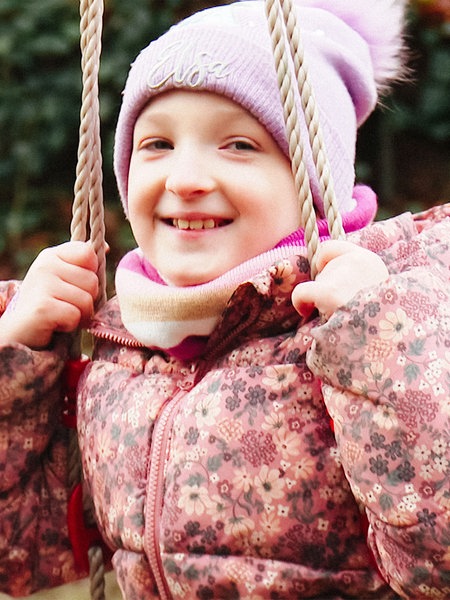 Melina, ein junges Mädchen in rosa Winterjacke und rosa Bommelmütze, sitzt auf einer Schaukel und lacht in die Kamera