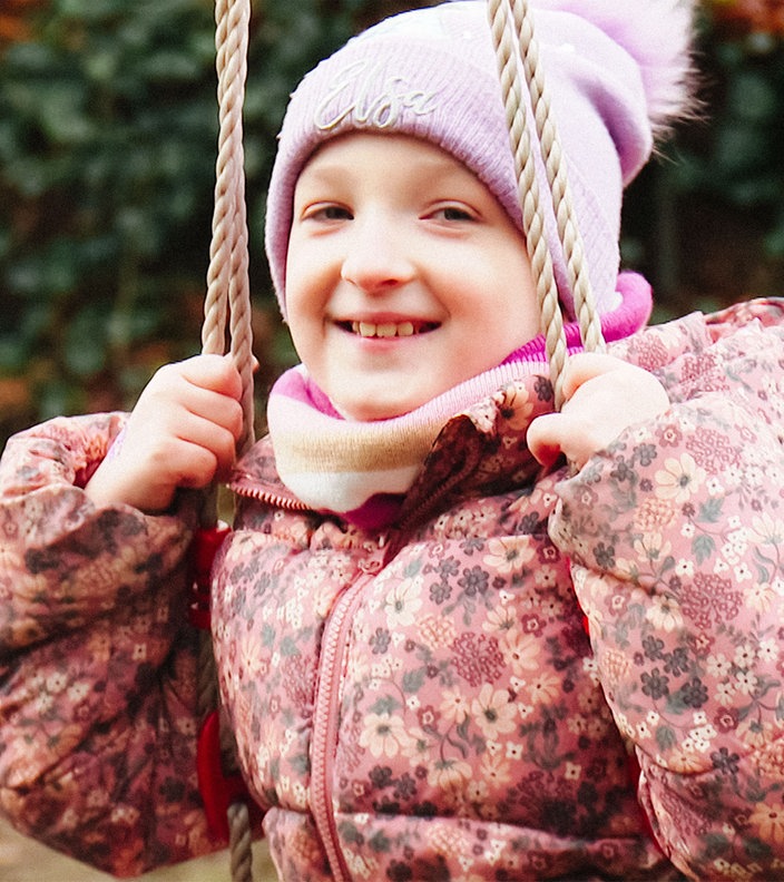 Melina, ein junges Mädchen in rosa Winterjacke und rosa Bommelmütze, sitzt auf einer Schaukel und lacht in die Kamera