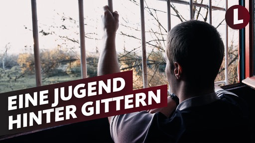 Ein junger Mann guckt von innen durch ein vergittertes Fenster nach draußen