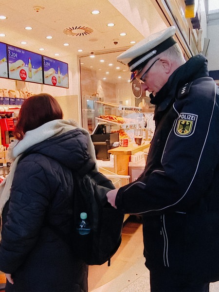 Ein Polizist steht hinter einer Frau, die in der Bahnhofsvorhalle in Münster Brötchen kauft. Er greift an ihren Rucksack.