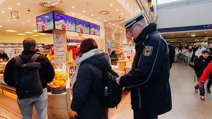 Ein Polizist steht hinter einer Frau, die in der Bahnhofsvorhalle in Münster Brötchen kauft. Er greift an ihren Rucksack.