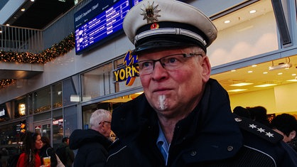 Jörg Schumacher im Portrait in der Bahnhofsvorhalle in Münster. Im Hintergrund ist eine Bäckerei sowie die Abfahrtstafel der Bahn abgebildet.