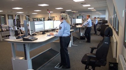 Blick in einen Büroraum in dem drei Personen in Polizeiuniform an Schreibtischen stehen und auf Monitore schauen