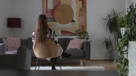 Eine Frau sitzt mit dem Rücken zur Kamera auf einem Stuhl in einem Wohnzimmer.