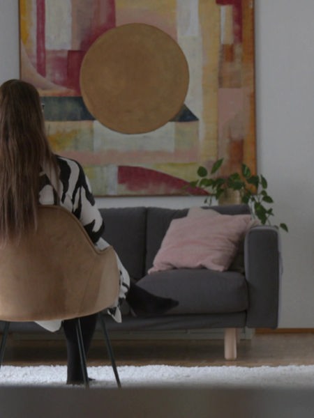 Eine Frau sitzt mit dem Rücken zur Kamera auf einem Stuhl in einem Wohnzimmer.
