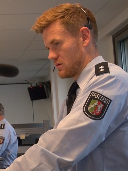 Mann mit rotblondem Haar und Bart in Polizeiuniform mit Headset auf dem Kopf schaut auf einen Computerbildschirm