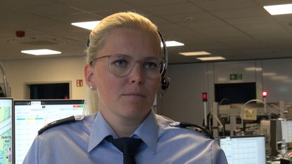 Frau mit hellblondem Haar, transparenter Brille in Polizeiuniform mit Headset auf den Kopf