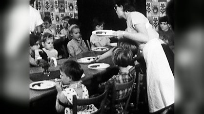 Archivbild. Kinder sitzen an Tischen und bekommen von Mitarbeiterinnen Teller mit Essen.