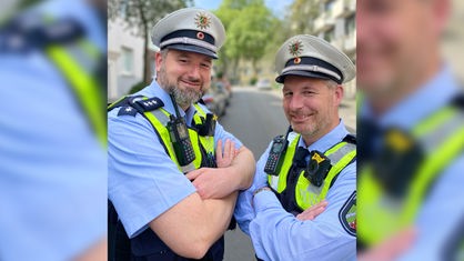 Man sieht zwei Polizisten mit Uniform, die mit verschränkten Armen in die Kamera lächeln