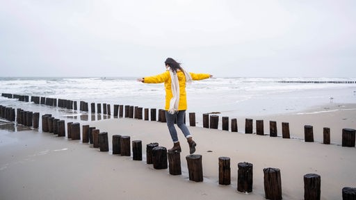 Eine Frau geht auf Holzpfählen am Strand entlang. Schriftzug auf dem Foto: "Grenzwertig. Ethik zwischen Leben und Tod"