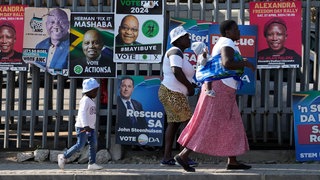 Das Beitragsbild des Dok5 Feature "Mandelas Erben - Südafrikas junge Generation ringt um die Zukunft" zeigt Frauen mit Kindern die an Wahlplakaten in Johannesburg vorbeigehen.