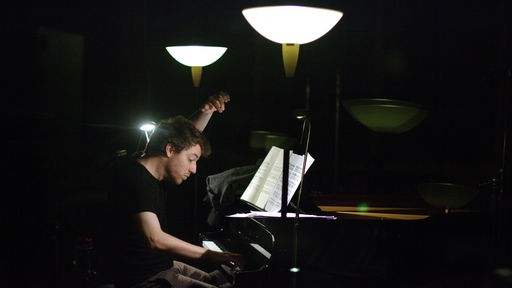 Der Pianist Cédric Pescia sitzt an einem schwarzen Flügel, der von zwei Lampen beleuchtet wird.