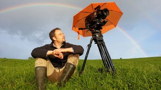 Jan Haft sitzt auf einer Wiese neben seinem Kamerastativ unter einem Regenbogen