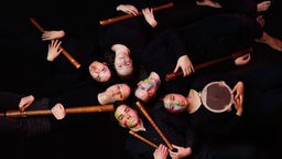 Draufsicht auf die liegenden Mitglieder des Flöten-Ensembles "Feuervogel" mit bunter Gesichtsbemalung und ihre Flöten in der Hand.